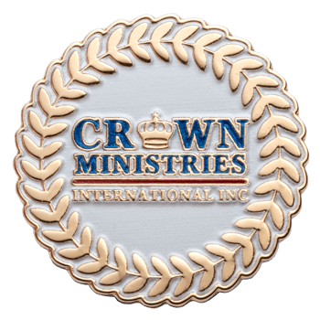 Round white and gold lapel pin for Crown Ministries International Inc with a crown emblem and laurel wreath border.