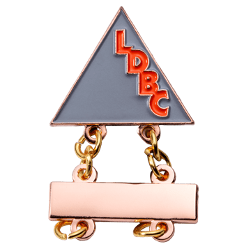 Triangular lapel pin with grey enamel and orange LDBC lettering, featuring a rose gold rectangular bar attachment joined by small chains.