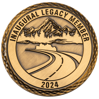 Antiqued gold challenge coin featuring a winding road leading toward mountains and evergreen trees with Inaugural Legacy Member 2024 text.