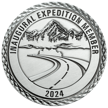 Antique silver challenge coin featuring a mountain range, pine trees, and a winding road with diamond-cut edges.